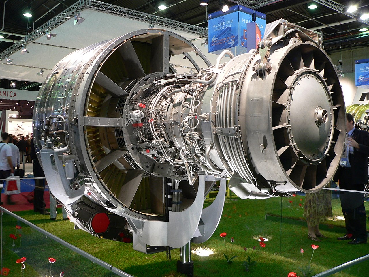 GE-Snecma CFM56-5B | An exposed jet engine at a trade show. The rear of the polished metal fan case is visible on the left. The outer casing of the compressor section, covered in fuel lines and electrical wires is to the right of the fan case. The right of the image shows the back of the engine, the exhaust area of the turbine section.